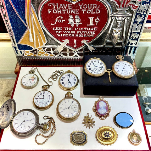 Several pocket watches and antique Victorian jewelry pieces in front of a vintage Mills Wizard Fortune Teller machine.