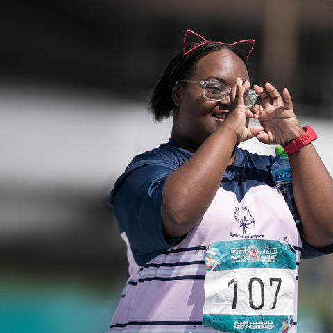 Auf dem Bild ist ein Mädchen zu sehen, das ein Trikot von den Weltspiele 2019 in Abu Dhabi trägt. Sie formt mit ihrem Hand ein Herz und hat ein breites Lächeln im Gesicht.