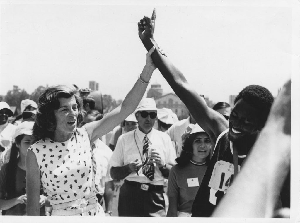 Eunice Kennedy Shriver und ein Athlet stehen nebeneinander, Kennedy Shriver hat den Arm des Athleten in der Hand und streckt ihn in die Höhe. Im Hintergrund sind weitere Menschen zu sehen.