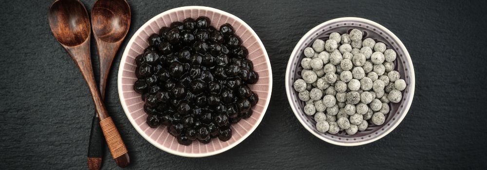 Uncooked tapioca pearls and brown sugar boba in bowls side by side with two wooden spoons 