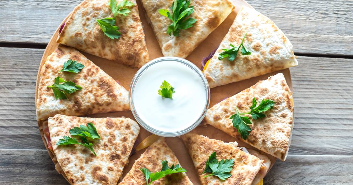 A circular wooden board holds eight quesadillas that surround a glass dish filled with sour cream topped with a garnish.