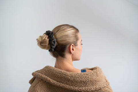 A blonde-haired model with her back to the camera looks over her shoulder with her hair up in a bun secured with a dark blue, plastic-free, organic cotton scrunchie.