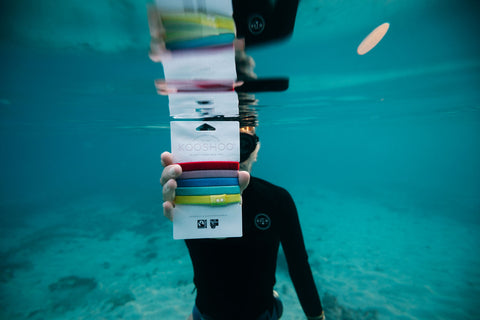 A model wearing a black rash guard holds up a pack of KOOSHOO plastic-free hair ties while underwater.