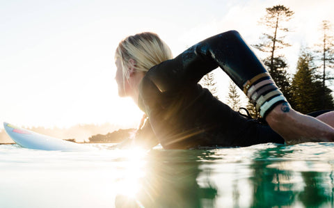 Aide paddles in the water on a surfboard wearing a black long-sleeved swim suit while her left arm is adorned with Kooshoo plastic-free hair ties.