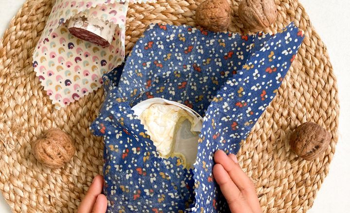 A woman in the process of wrapping cheese with a blue beeswax wrap. A half cured sausage is already wrapped next to it.