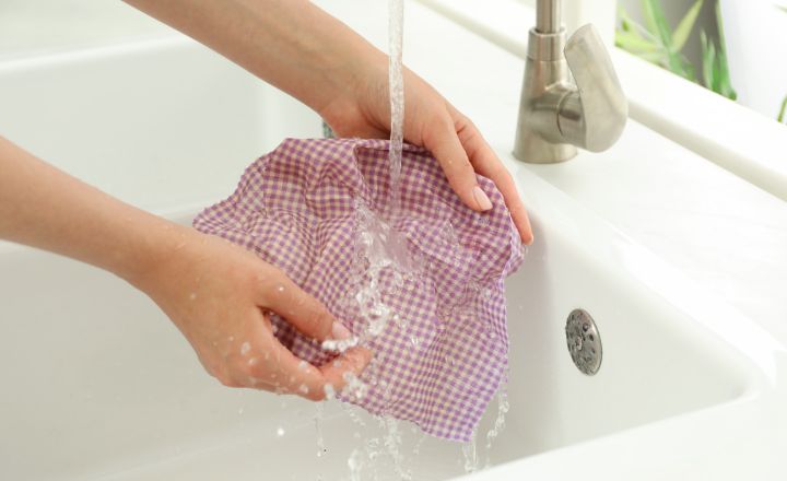 a woman holding a single beeswax wrap in the sink, under running water