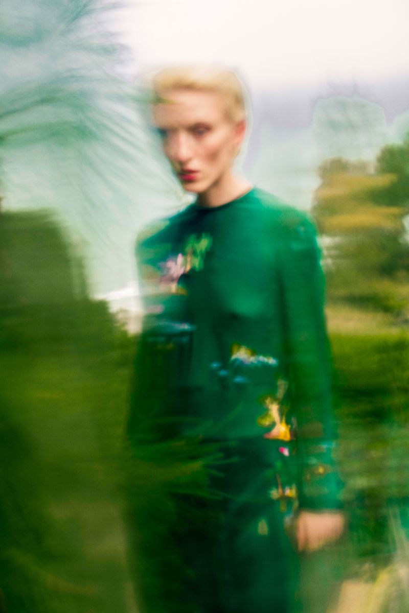 Blurry photograph of woman in green dress with blonde hair amongst green trees