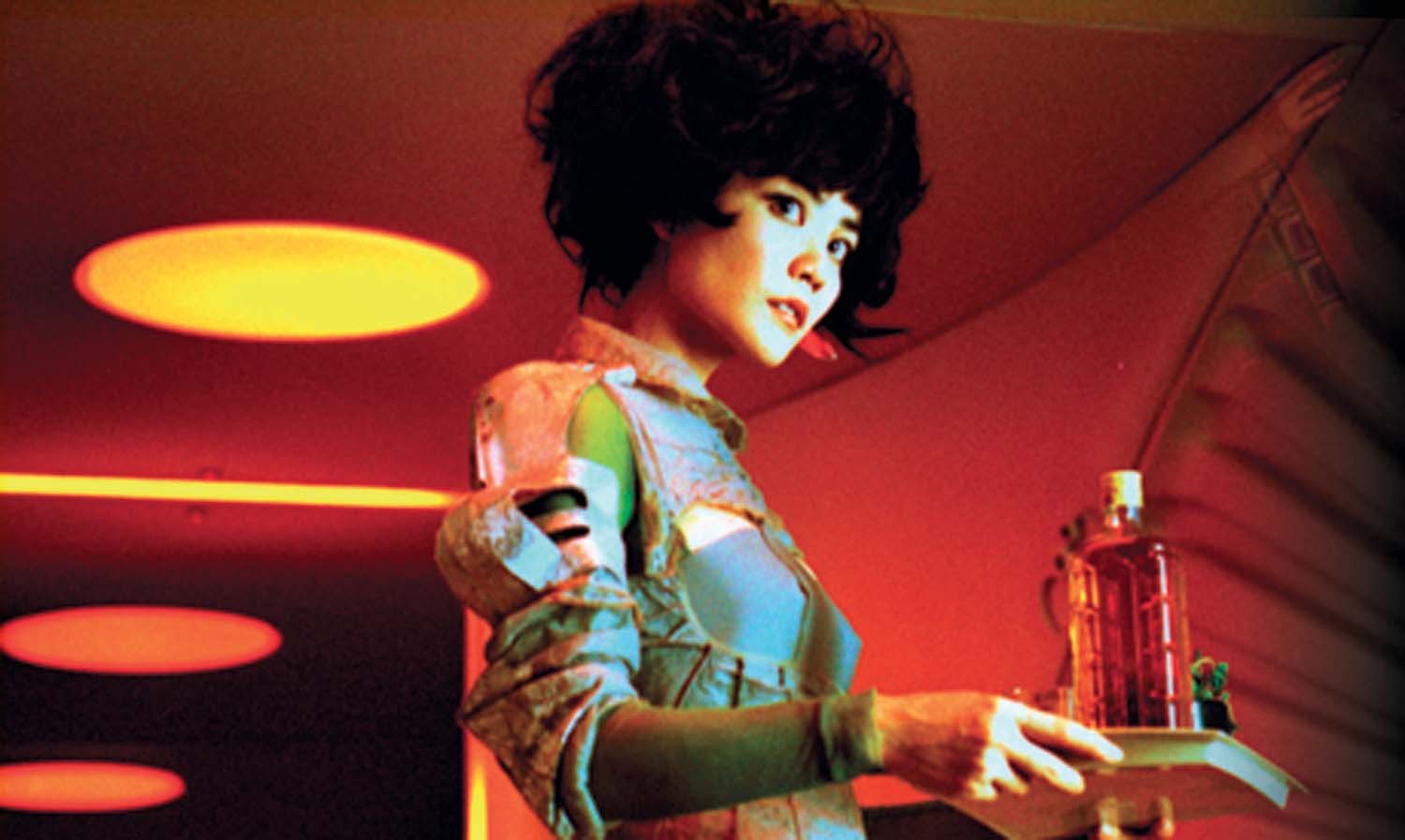 Still from Wong Kar-wai's film 2046 showing a woman with dark bouffant hairstyle and a retro futurist style dress looking offscreen and carrying a tray of drinks