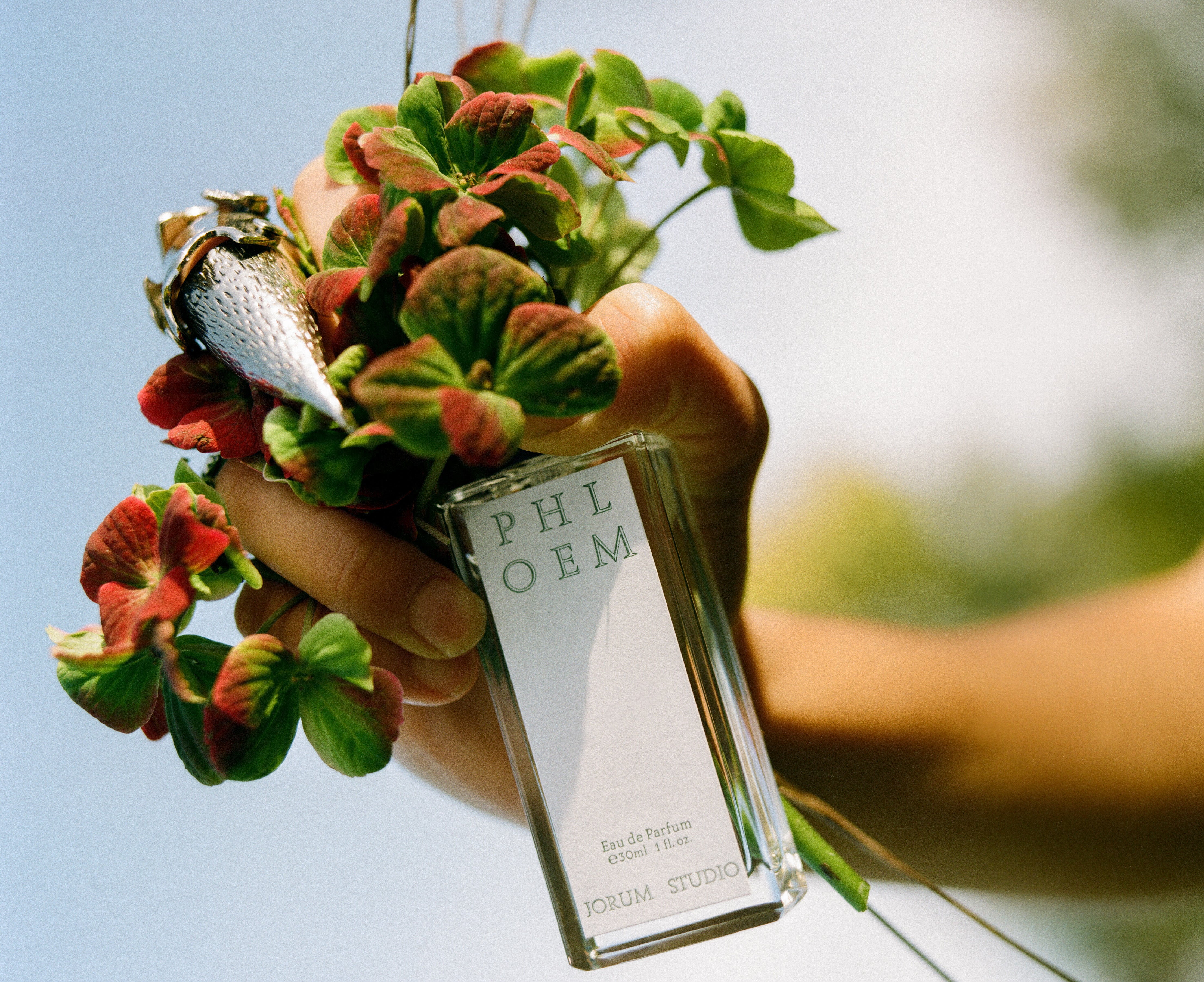 hand with pointed metal claw ring holding Jorum Studio Phloem perfume bottle and flowers photograph by Craig McIntosh