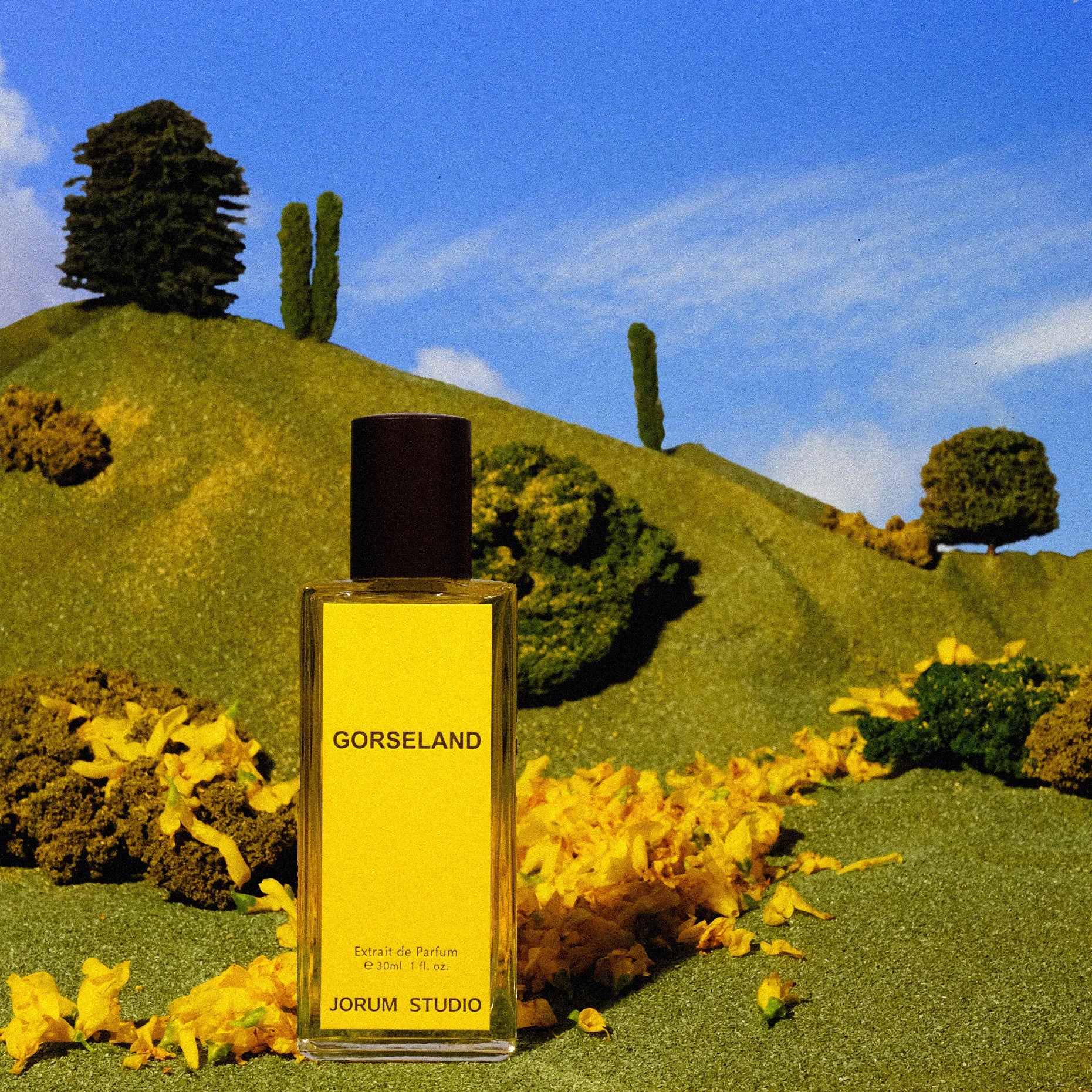 Bottle of Jorum Studio Gorseland perfume superimposed on a small scale model hill with model trees and scattered gorse petals