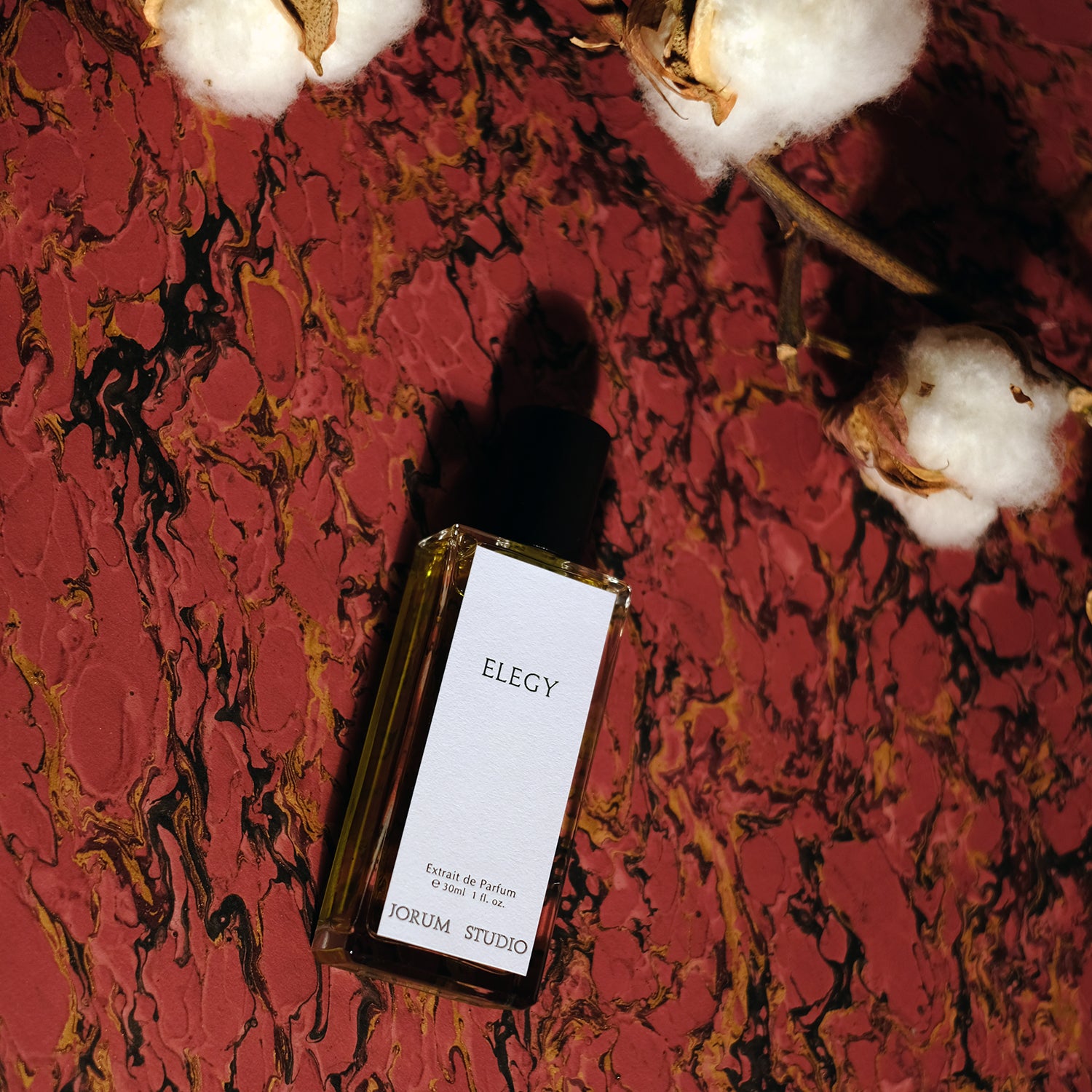 Bottle of Jorum Studio Elegy perfume with cotton flowers and red marble background