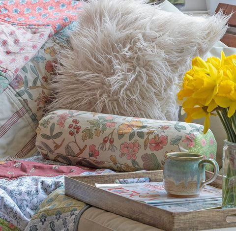 a gloral yoga pillow positioned on a sofa, next to a fluffy cushion and in front of a coffee table that's home to a book and some yellow flowers in a vase