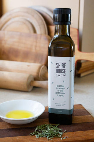 A bottle of olive oil next to an oil filled dish and some rosemary leaves in front. Resting on a wooden chopping board with chopping boards behind