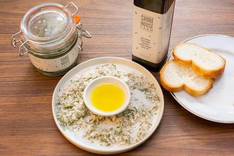 A ceramic dukkah dish filled with salt and oil. Surrounded by salt jar, olive oil bottle and a plate of bread, all resting on a wooden surface