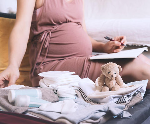 Pregnant women writing something in a notebook and touching her baby clothes