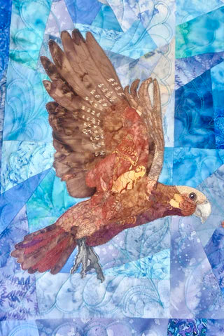 A patchwork quilt featuring a brown kea bird flying across blues and purples patchworked together by textile artist Sue Duffy