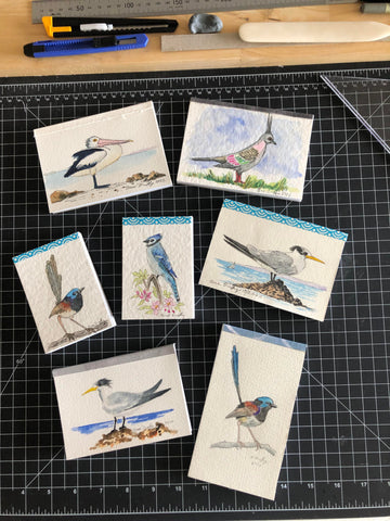 hand-made note pads with watercolour paintings of birds layed out on a cutting mat in artist studio of Sue Duffy featuring a wren, tern, pelican, and native pigeon
