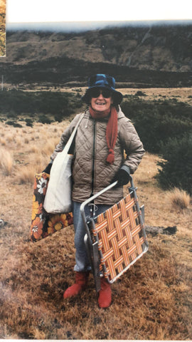 little old lady artist  with a hat and sunglasses wearing a puffer jacket and gloves standing in nature holding a deck chair and two bags of art supplies to do some plein aire painting in Cambridge New Zealand