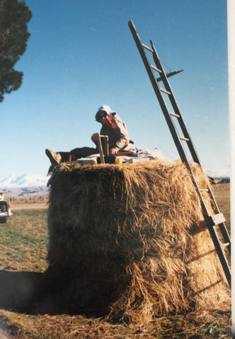 old lady artist sitting on a haystack in the south of New Zealand with her plein aire paint set up a ladder leans against a tall hay bail as she organises her paints