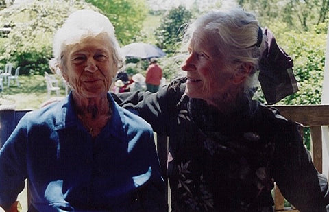 two little old ladies with white hair one wearing a scarf tied in her hair the other a blue collared dress on a veranda in the half sun looking out onto a garden with tables