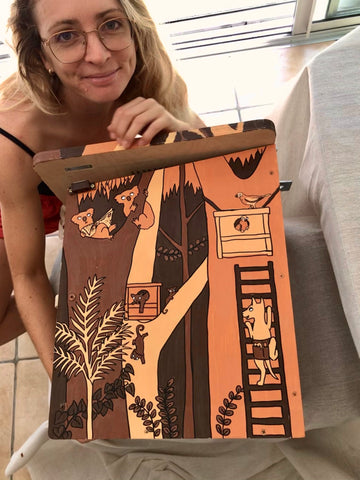 An artist with her artwork a painted nest box, the painting is of a dingo climbing a ladder to a nest box in the Australian eucalypt forest and some koalas are reading the newspaper watching as some ringtail possums play on a nest box in the background.