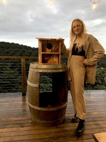 Artist standing next to her painted nest box selling for Auction. Nest box sits on top of a barrel overlooking rainforest at Byron Bay's Brookie's Gin