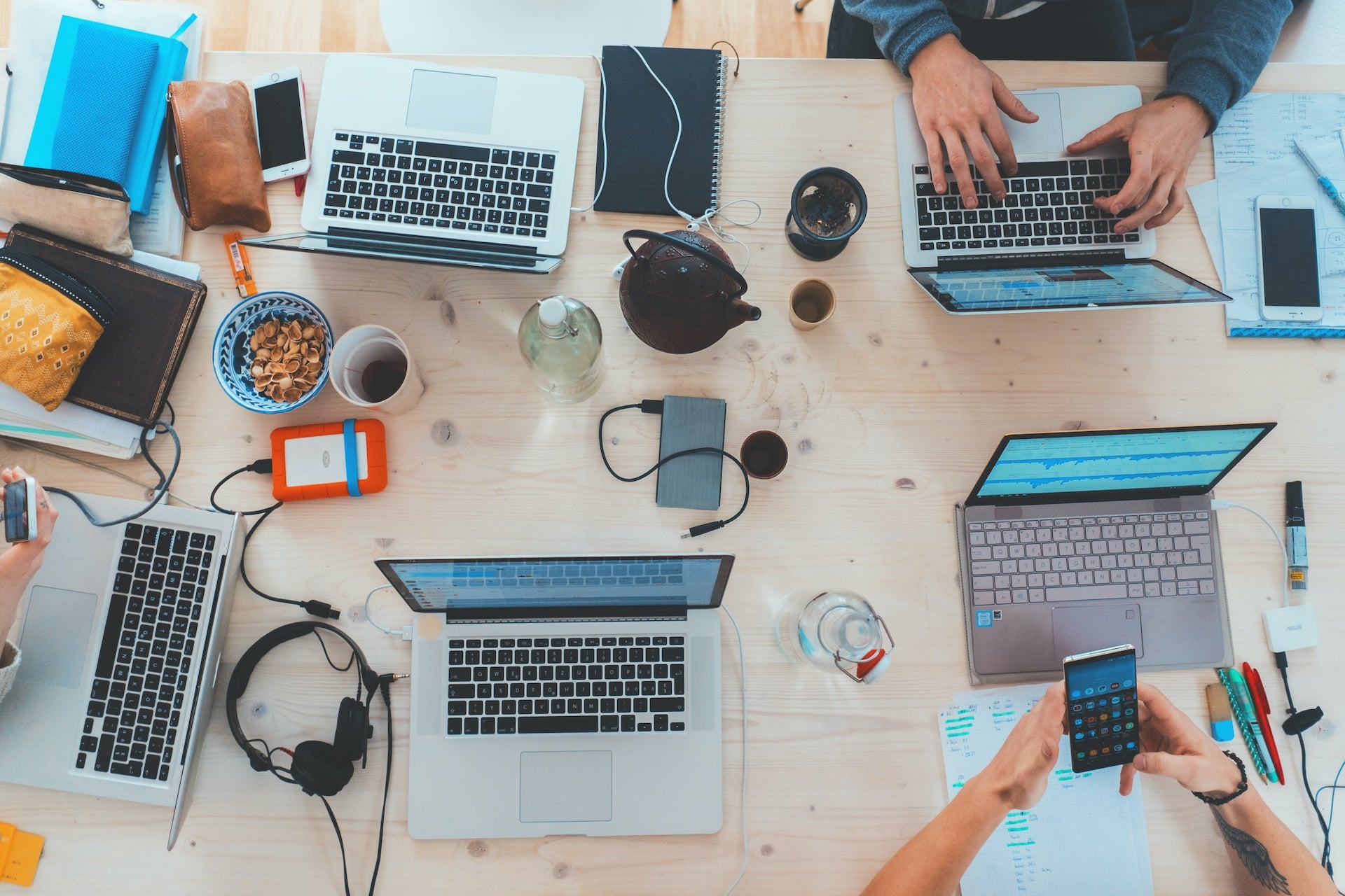 Vista aérea de una mesa de madera con cuatro personas trabajando en computadoras portátiles, junto con artículos personales como teléfonos, computadoras portátiles y una tetera. Hay una sensación relajada y colaborativa en el espacio.