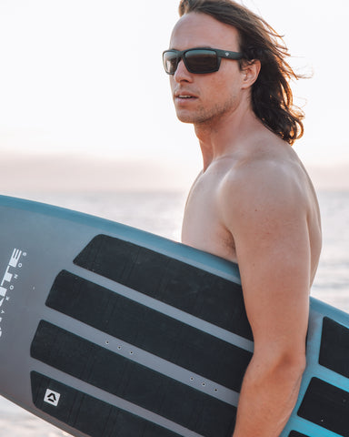 a man holding a surfboard on the beach wearing Torege watersports sunglasses