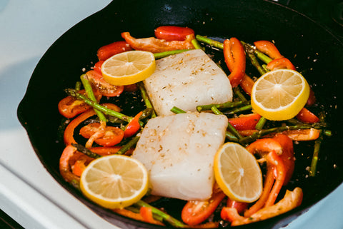 two portions of wild cod in a cast iron skillet with vegetables and lemons surrounding it