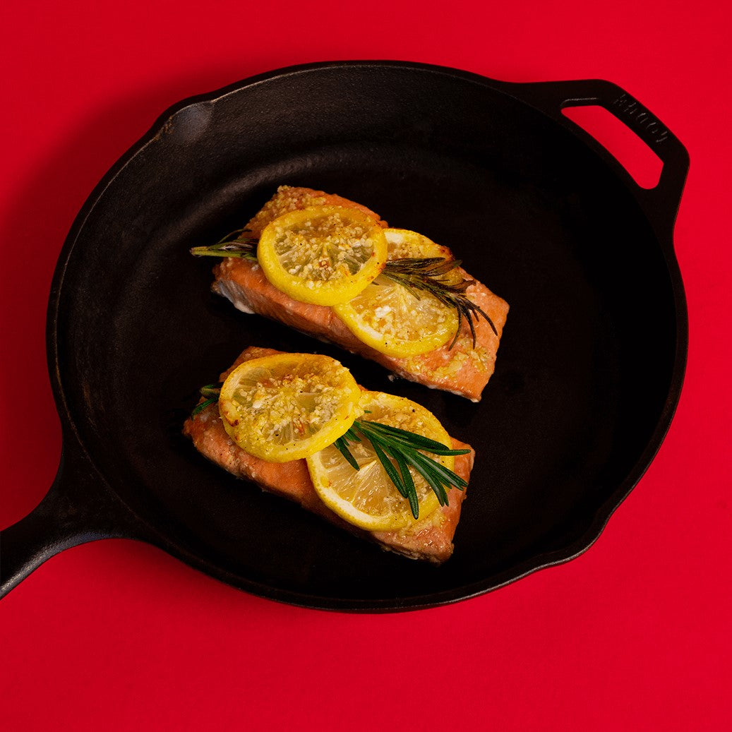 Roasted wild salmon topped with lemon and rosemary in a cast iron pan on a red background