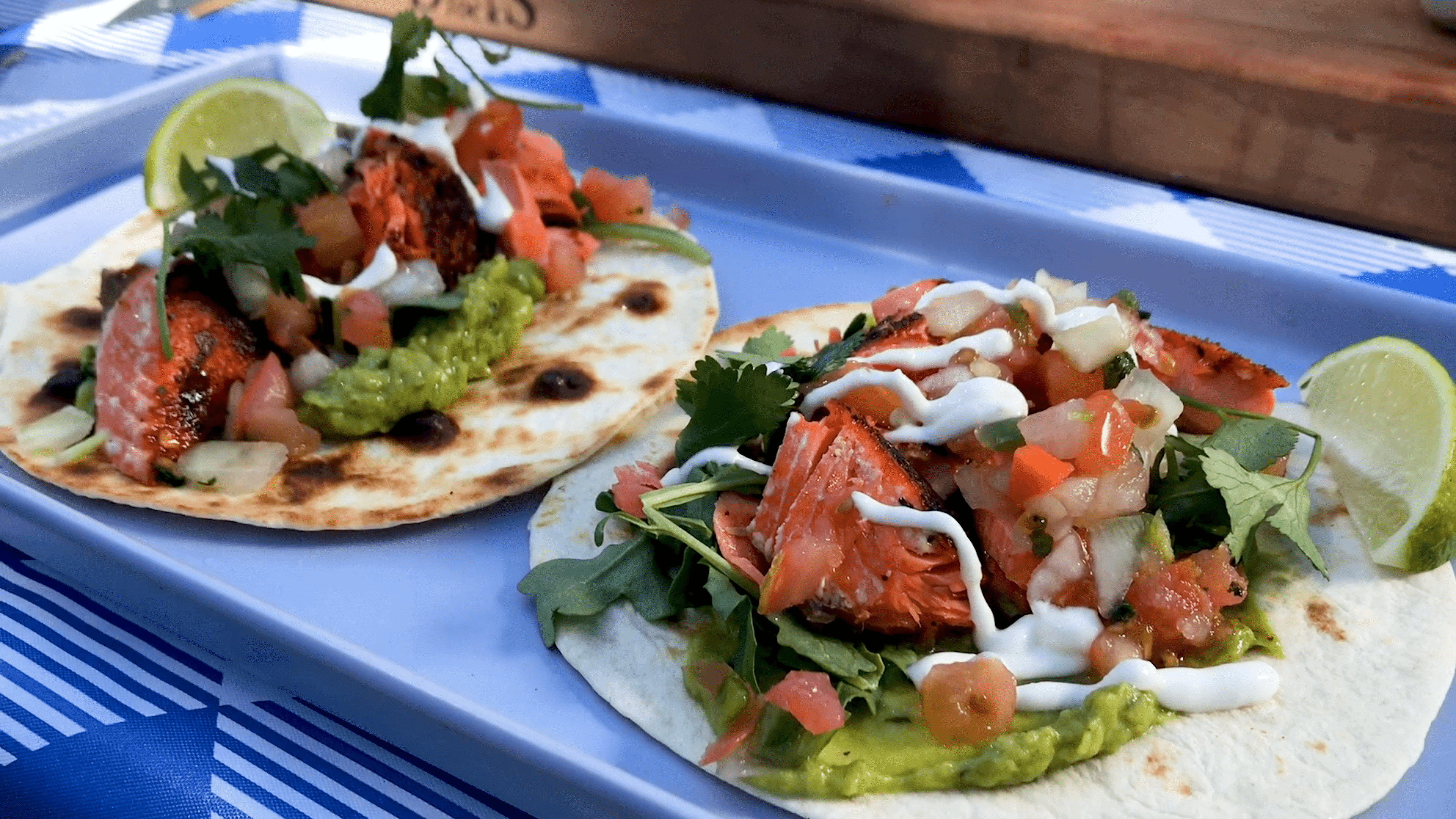 two prepared salmon tacos on tortillas with guacamole, salsa, arugula, cilantro, and sour cream with lime wedges on the side on a plate on a picnic table