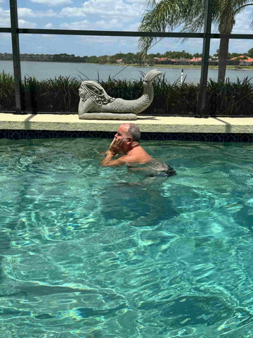 John Milan poses in a swimming pool, mimicking a mermaid statue behind him.