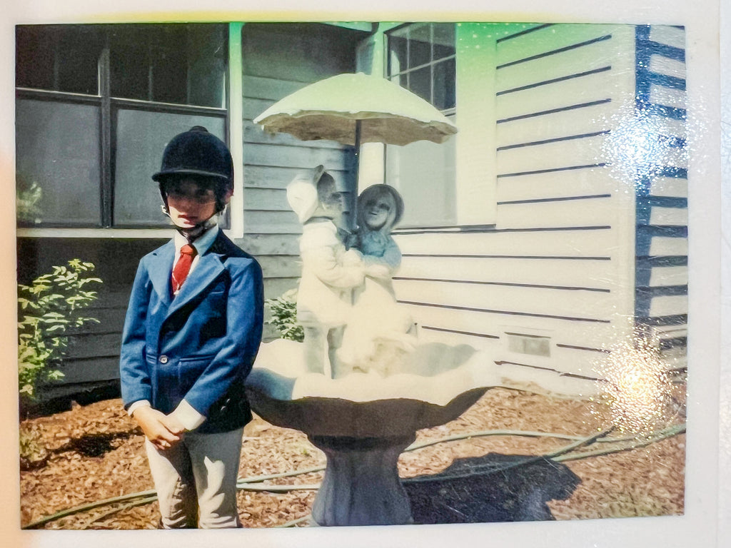 Elli Milan as a little girl, dressed in her equestrain gear, standing and smiling in front of a fountain.