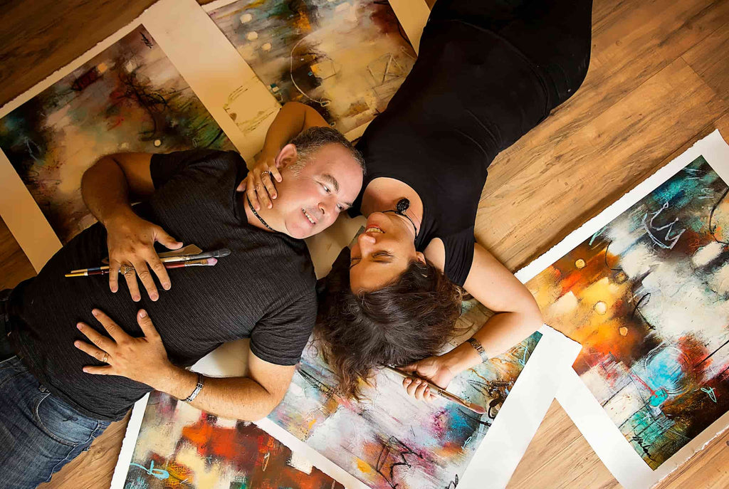 John and Elli Milan gaze at each other while lying on top of their collaborative artwork and gripping paintbrushes