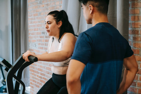 White female on an exercise bike in a crop top as trainer watches