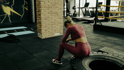 Female in coral crop top and leggings sitting on a tyre in a gym with her head down