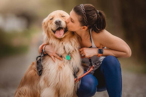 Woman in blue leggings and tank top kneels down and kisses her happy Golden Retriever on snout