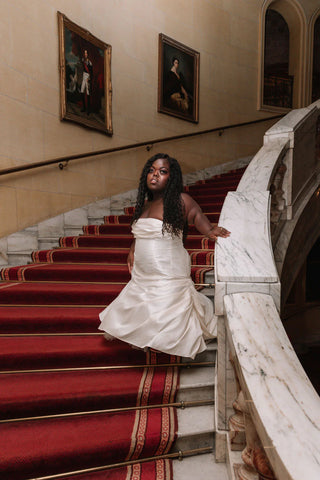 A woman with a deep skin tone and dark hair is positioned on a staircase, wearing a custom-made white wedding dress. The dress has an A-line silhouette, a bandeau neckline, and a ruffled hem.