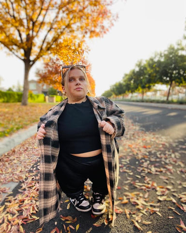 Jonna poses in a park with an autumnal ambience, wearing a black t-shirt and black leggings paired with Nike trainers in black and white. Completing her outfit, she adds a checkered black and beige unbuttoned jacket and stylishly places sunglasses atop her head.