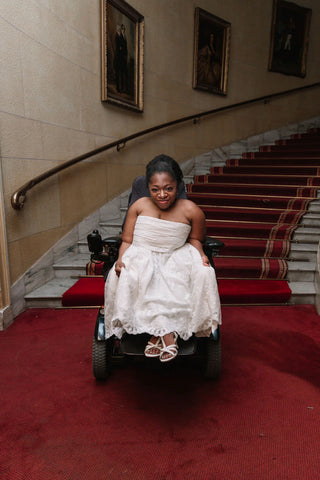 On a staircase, a woman with a deep skin tone and dark hair sits in a wheelchair. She is elegantly dressed in a custom-made white wedding gown. The dress features an A-line silhouette and a bandeau neckline, while the bottom half of the dress is adorned with intricate lace detailing that extends all the way down to the hem.