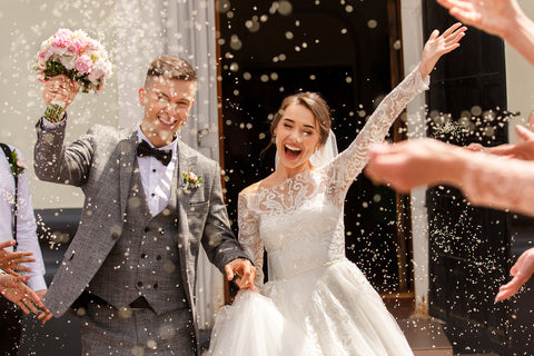 A couple embraces, holding hands amidst confetti showering down on them. The groom grasps the bride's pink bouquet, while the bride joyfully raises her arm in celebration.