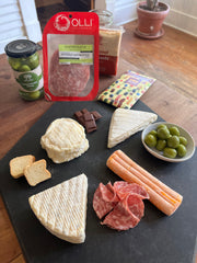 Display of 4 cheeses, olives, sliced meat, chocolate and crackers on black cheese board