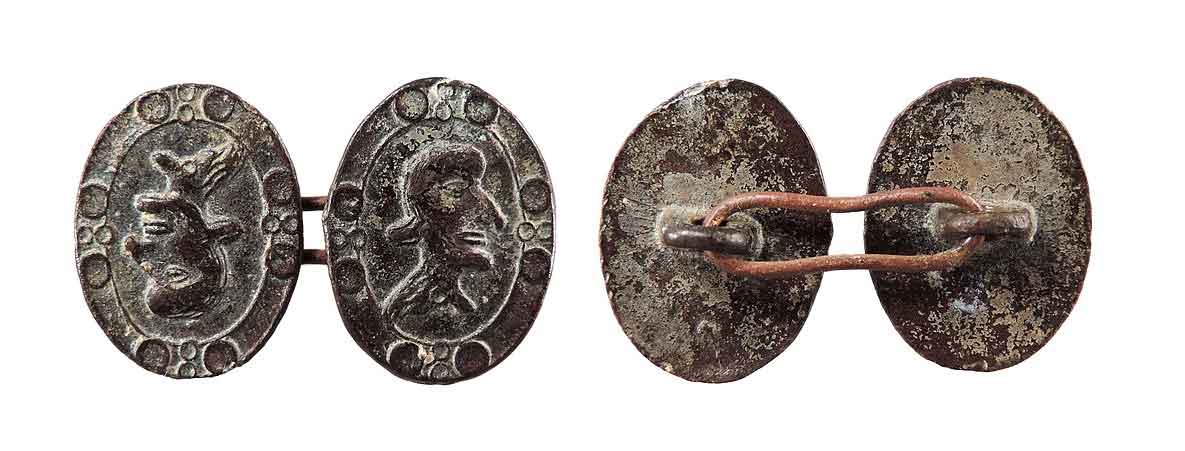 A weathered cuff link made of two metal discs stamped with an image of a man's head with a large nose