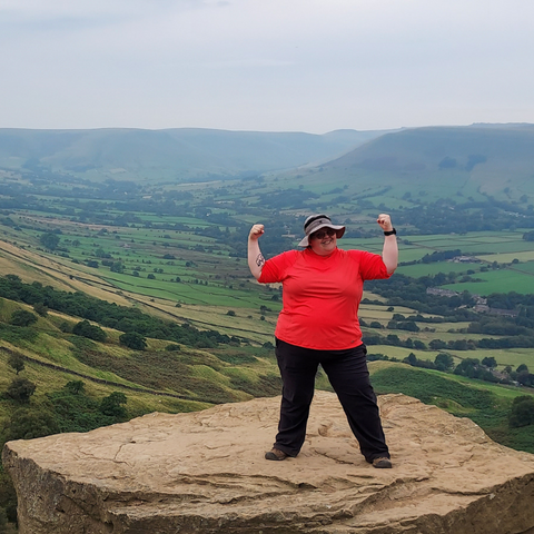fat woman in red top and black trousers stands triumphantly on a rock with the valley below her