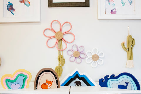 four wood toy puzzles on a shelf, they are shaped as animals with stacking pieces that resemble where the animals lives. then macrame pieces shaped as flowers and a cactushanging on the wall above them