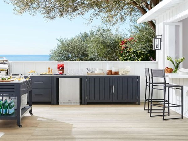 Front view of a 5.12 Cu Ft Undercounter Beverage Outdoor Refrigerator installed beneath the kitchen table, surrounded by various kitchen appliances in an outdoor kitchen setting. In the background, a picturesque beach scene is visible, with a few trees adding to the serene atmosphere