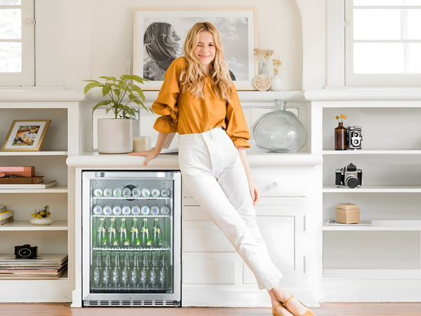 Front view of a living room setup with a woman standing in the middle, smiling and leaning against the 5.12 Cu Ft Beverage Outdoor Refrigerator 132 cans installed beneath a table, surrounded by decorations