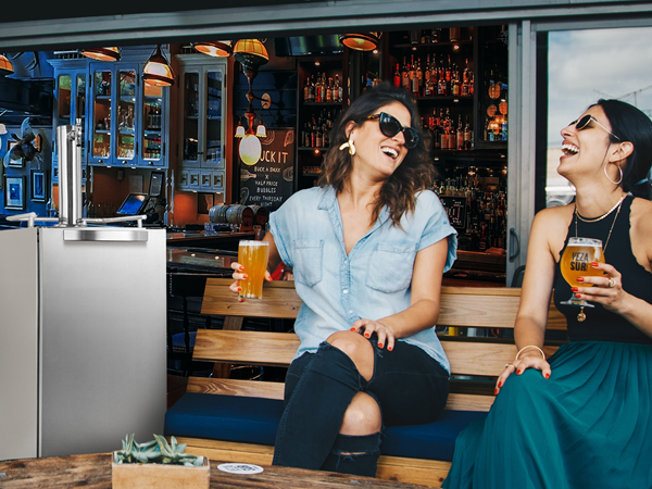 A 3.2 Cu Ft Outdoor Refrigerator Kegerator with a capacity of 96 cans is positioned at the left corner in a bar setting. Two women are seated beside the product, smiling at each other.
