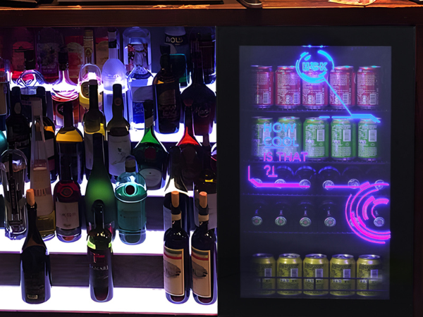 Front view of the 3.5 Cu Ft Cyberpunk Glass Door Beverage Fridge installed under the bar counter, positioned next to a wine display shelf