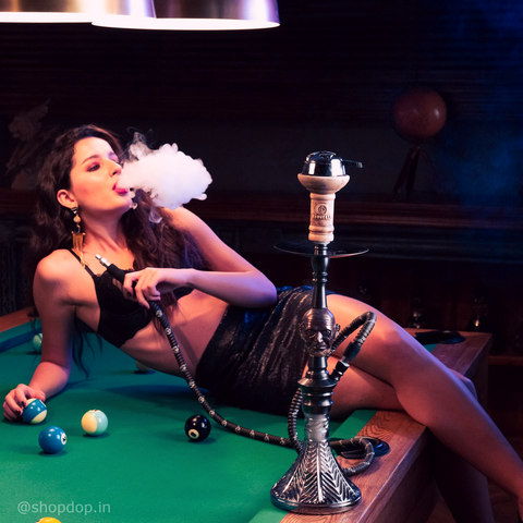 In this image you can see Cocoyaya Dr. D Hookah Bohemian Series with a female model smoking out from the hookah and using HMD device over chillum. She is lying over on a snooker table. 
